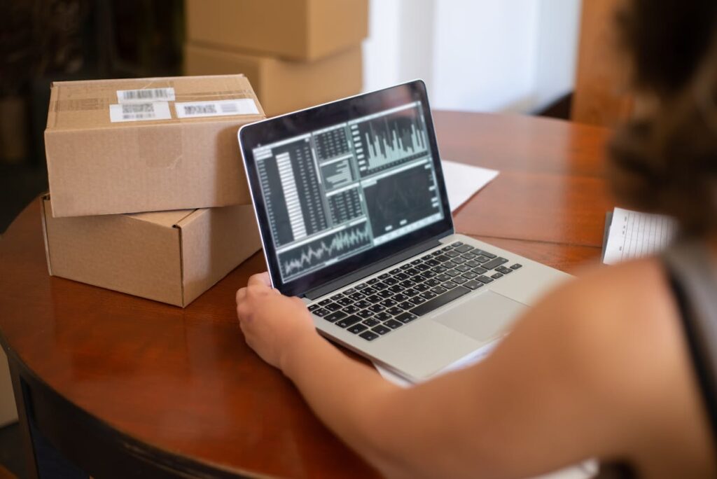 Close-up image of a woman managing analytics on a laptop beside shipping boxes at a table showing single mom struggling financially. 