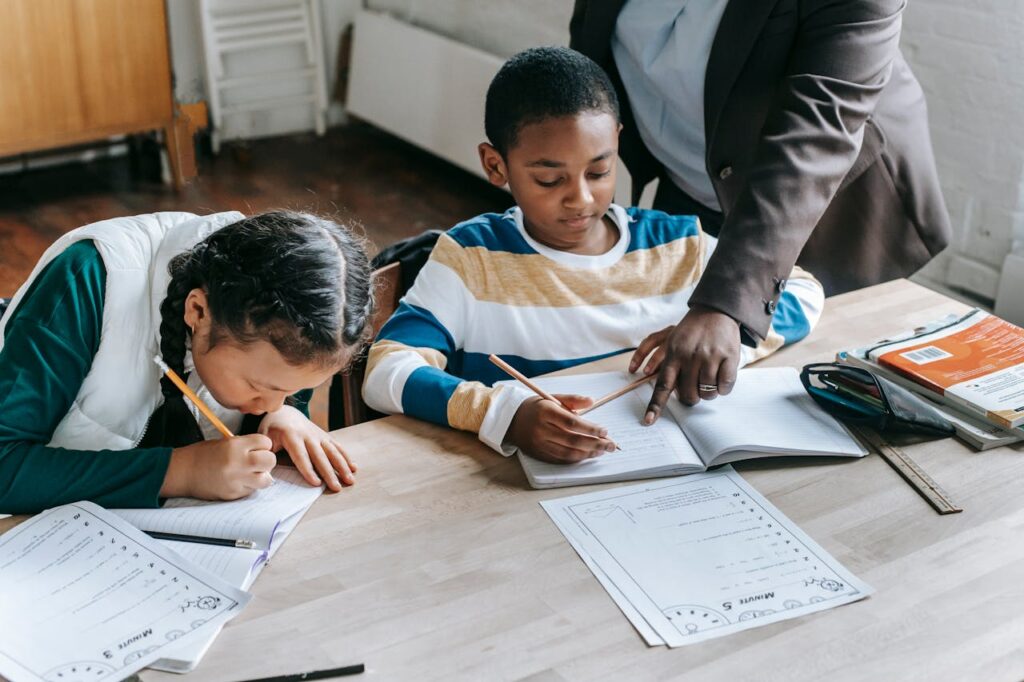 Image of two children writing in notebooks with a teacher assisting them at a wooden table representing Office Time of a WFH Mom.