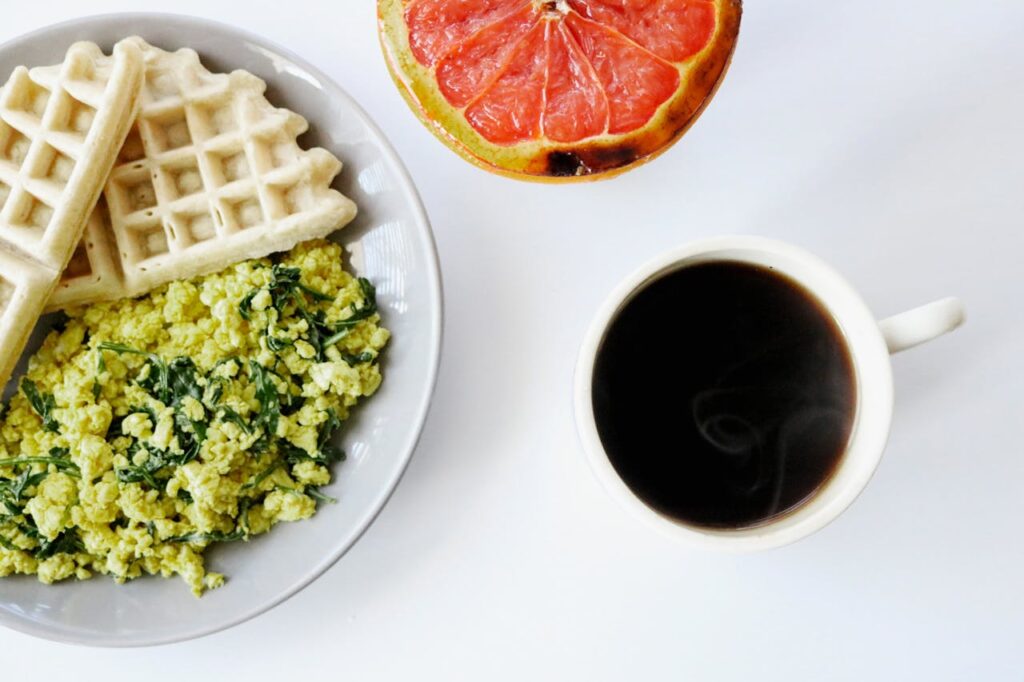 Close-up of breakfast plate featuring scrambled eggs mixed with greens, two waffle pieces, a half-cut grapefruit, and a cup of black coffee, all arranged on a white surface.