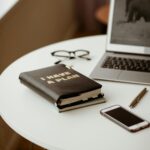 Image of a workspace featuring a white round table. On the table, there is a black notebook with gold lettering that reads "I HAVE A PLAN", a pair of black-framed glasses, a silver pen, and a smartphone with a black screen. A partially visible laptop with an open screen displaying a blurred image is also present. The scene is well-lit, evoking a calm and organized atmosphere showing Couple Vision Board.