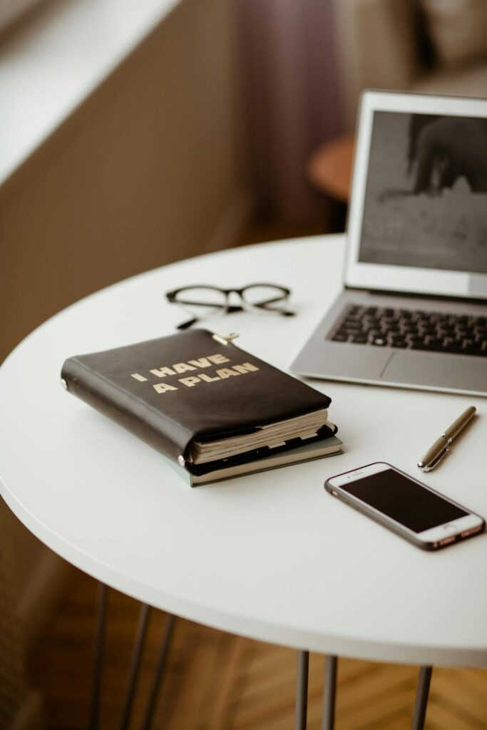 Image of a workspace featuring a white round table. On the table, there is a black notebook with gold lettering that reads "I HAVE A PLAN", a pair of black-framed glasses, a silver pen, and a smartphone with a black screen. A partially visible laptop with an open screen displaying a blurred image is also present. The scene is well-lit, evoking a calm and organized atmosphere showing Couple Vision Board.