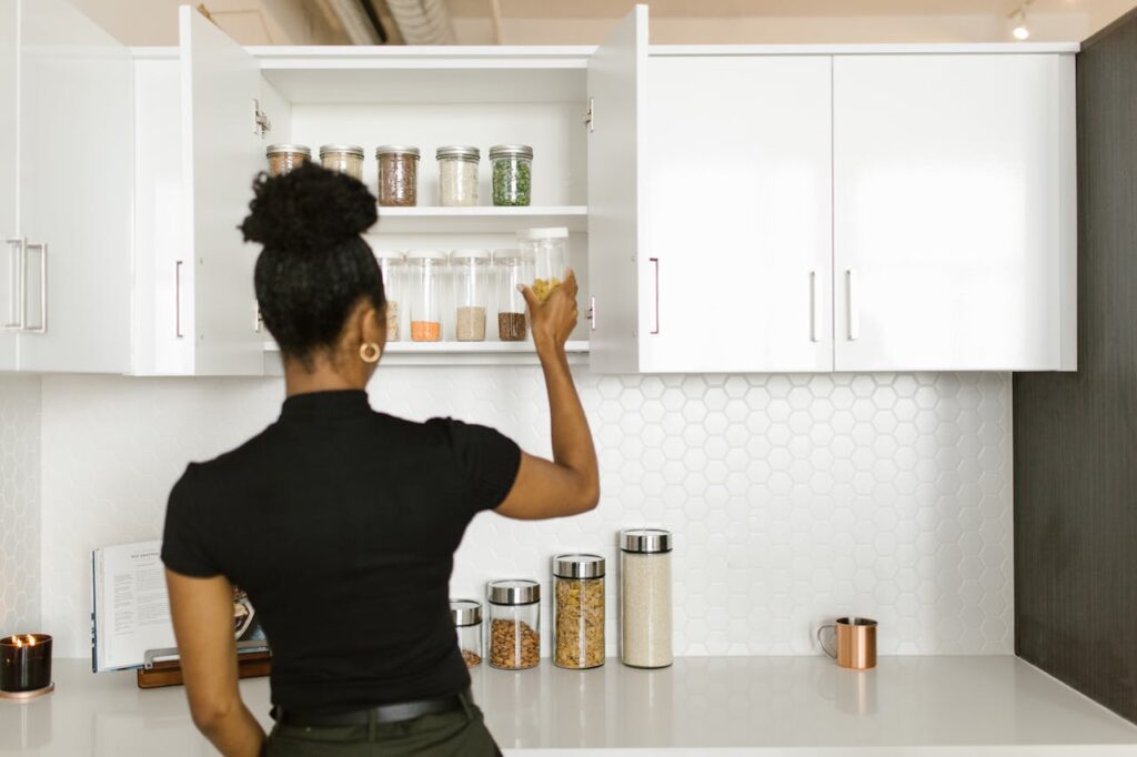 Close-up image of a woman organizing jars of dry goods in a white kitchen cabinet, with additional labeled jars neatly arranged on the countertop below, set against a hexagonal tile backsplash representing meal planning for busy families.