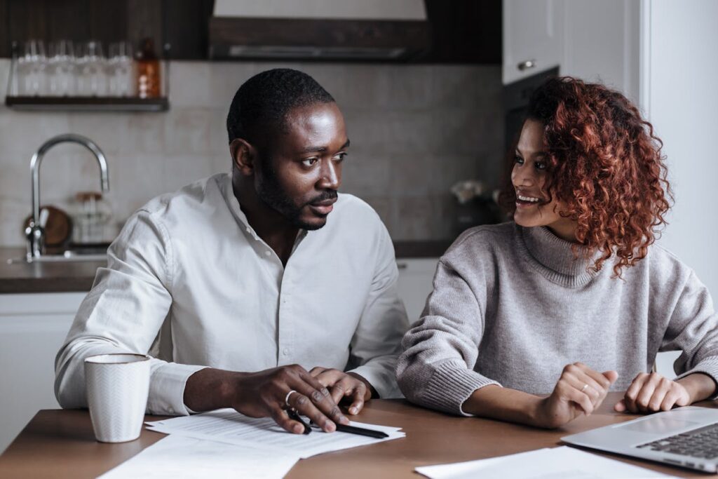 Image of a man and a woman are sitting at a table, engaged in a discussion. The man, wearing a light-colored shirt, is leaning slightly forward with his hands on some documents, looking attentive. The woman, wearing a cozy gray sweater, is smiling as she looks at him. A laptop and coffee mug are on the table, suggesting they are working or collaborating in a casual home or office setting. The background features a kitchen with a modern design, including a sink and shelves with glasses, representing couple dream board.