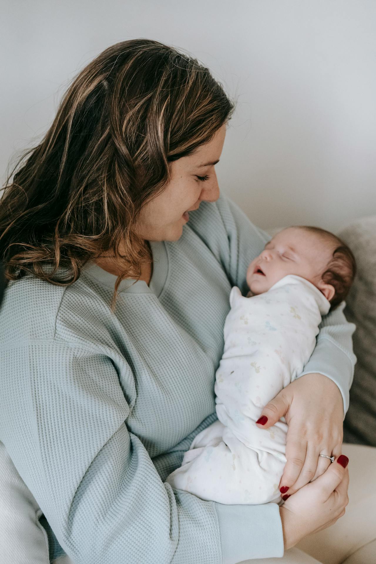 Image of a smiling woman in a light sweater holding a sleeping newborn wrapped in a white blanket representing 5 Days Around the Bed.