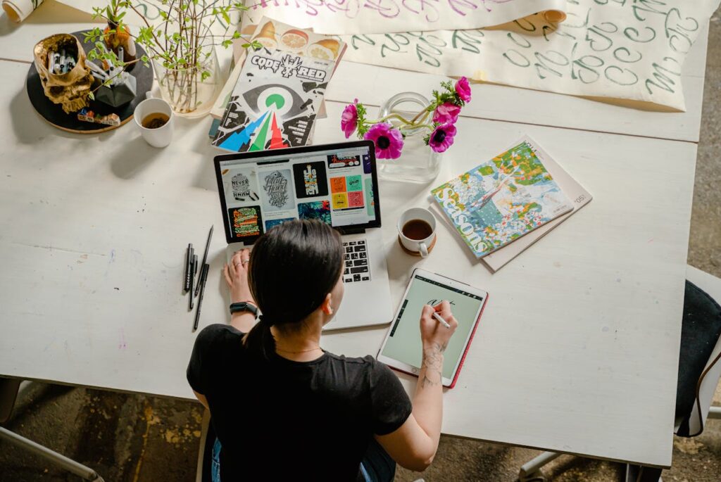 Image of a woman working on creative design at a desk with a laptop, tablet, flowers, and art materials showing side hustle for single moms online.