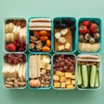 Image of an array of colorful snack boxes filled with various healthy foods, including crackers, nuts, berries, cheese, boiled eggs, cucumber sticks, grapes, cherry tomatoes, and sandwiches, arranged neatly on a light green background. Image used for the article Meal Planning For Working Moms.