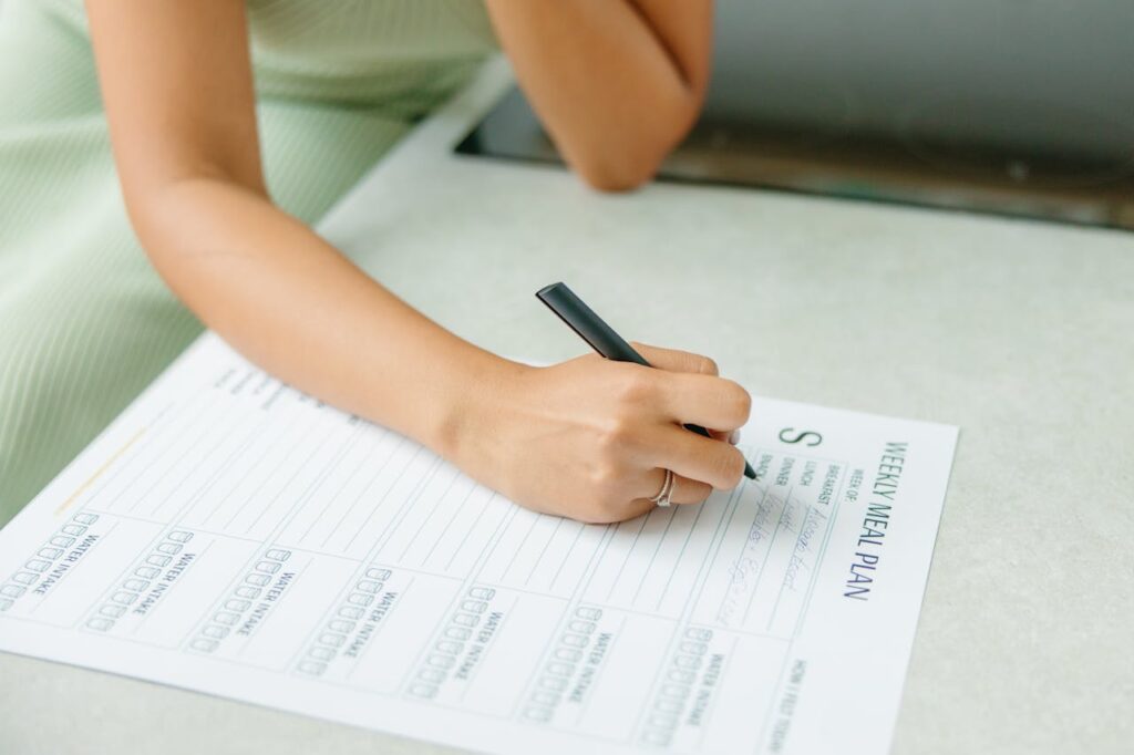 Image of a person writing on a Weekly Meal Plan sheet with a black pen. The focus is on their hand and the paper, with a blurred background.