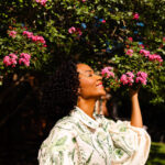 Image of a woman with curly hair wearing a floral shirt enjoys the scent of pink blossoms on a tree, showing how to feel like yourself again.