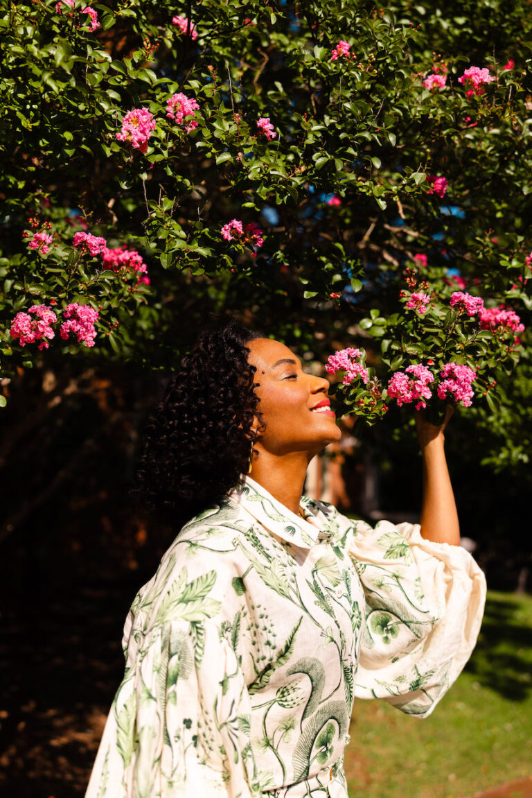 Image of a woman with curly hair wearing a floral shirt enjoys the scent of pink blossoms on a tree, showing how to feel like yourself again.