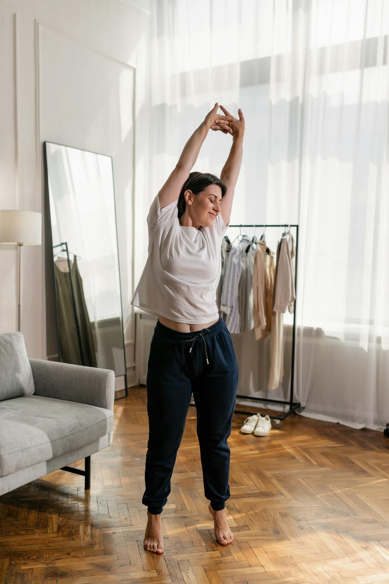 Image of a relaxed woman in casual clothes stretching in a bright room with wooden flooring and natural light.