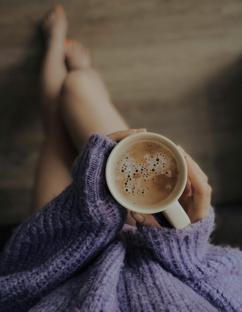 Image of a woman holding a cup of coffee while wearing a cozy purple sweater, legs crossed, and relaxed.