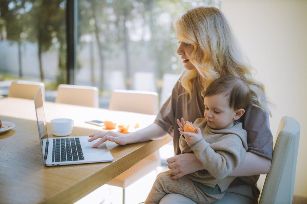 Image of a woman with blonde hair working on a laptop while holding a toddler eating an orange slice, representing how to be a good working mom.