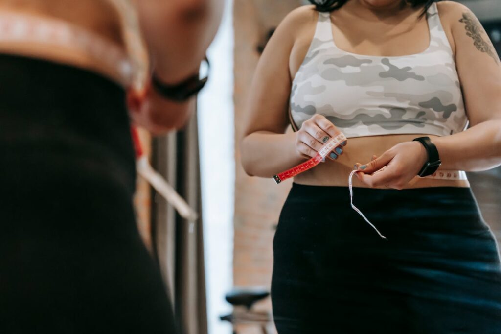 Image of a woman measuring her waist with a tape while wearing workout attire in front of a mirror, representing how can I dress stylishly as a plus-size mom without feeling self-conscious.