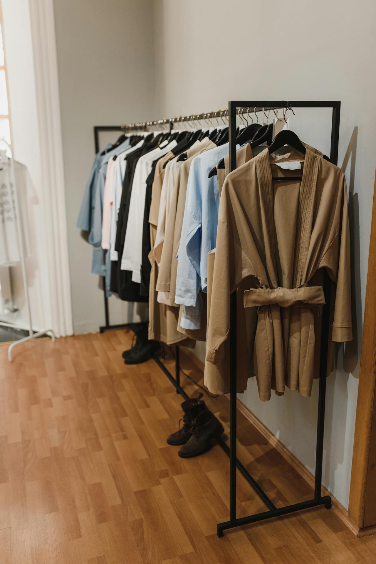 Image of a minimalist clothing rack displays neutral-toned garments and black boots on a wooden floor, representing build an eco-friendly wardrobe as a mom.