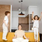 Image of a woman meditating on a yellow couch while two children jump energetically around her, showing Struggles of a Stay-at-Home Mom.