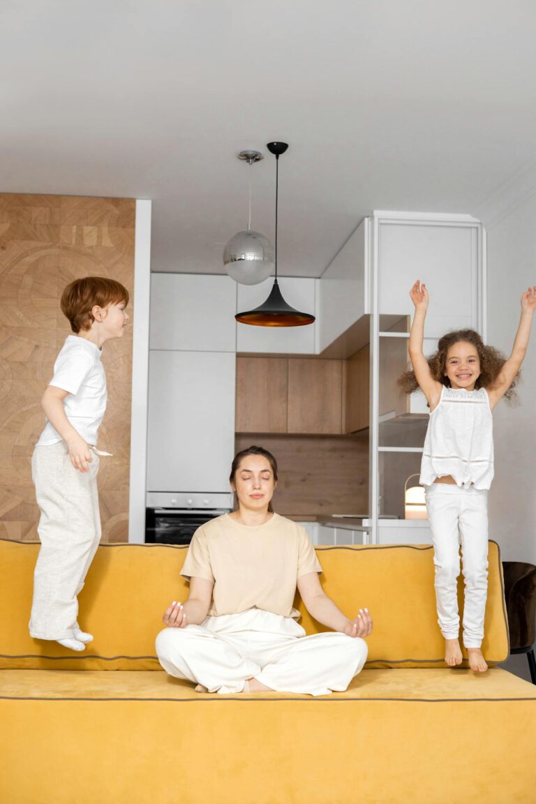 Image of a woman meditating on a yellow couch while two children jump energetically around her, showing Struggles of a Stay-at-Home Mom.