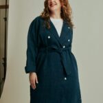 Image of a smiling woman with curly red hair in a double-breasted navy coat, standing in a studio, showing how can I dress stylishly as a plus-size mom without feeling self-conscious.