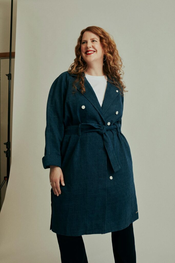 Image of a smiling woman with curly red hair in a double-breasted navy coat, standing in a studio, showing how can I dress stylishly as a plus-size mom without feeling self-conscious.