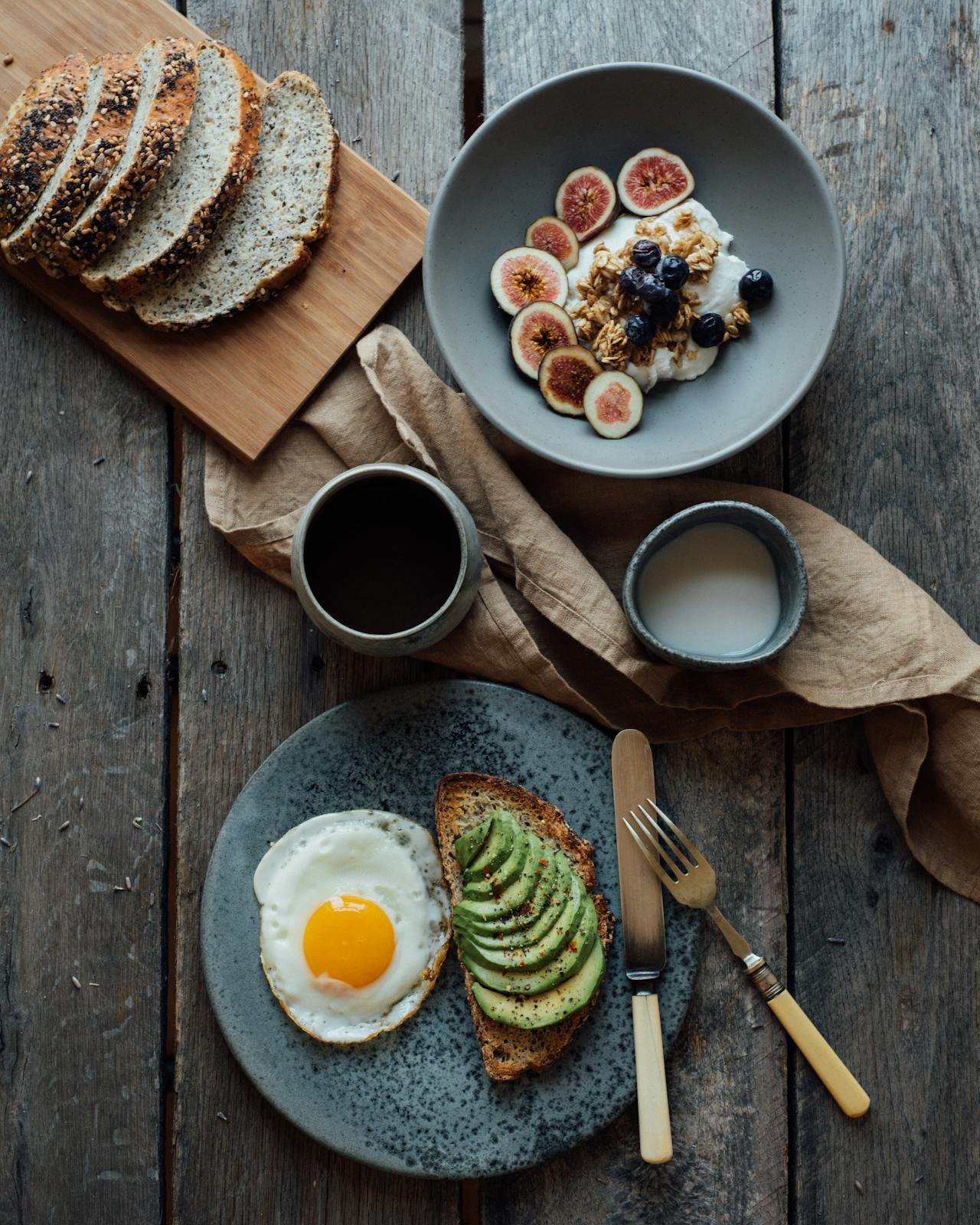 Image of breakfast spread with avocado toast, fried egg, sliced bread, and a bowl of yogurt topped with figs and berries.