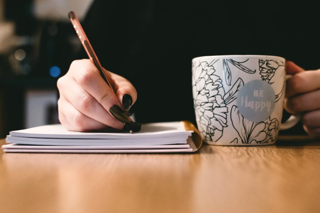 Close-up of a woman writing in a notebook while holding a mug that says 'Be Happy' on a wooden table.