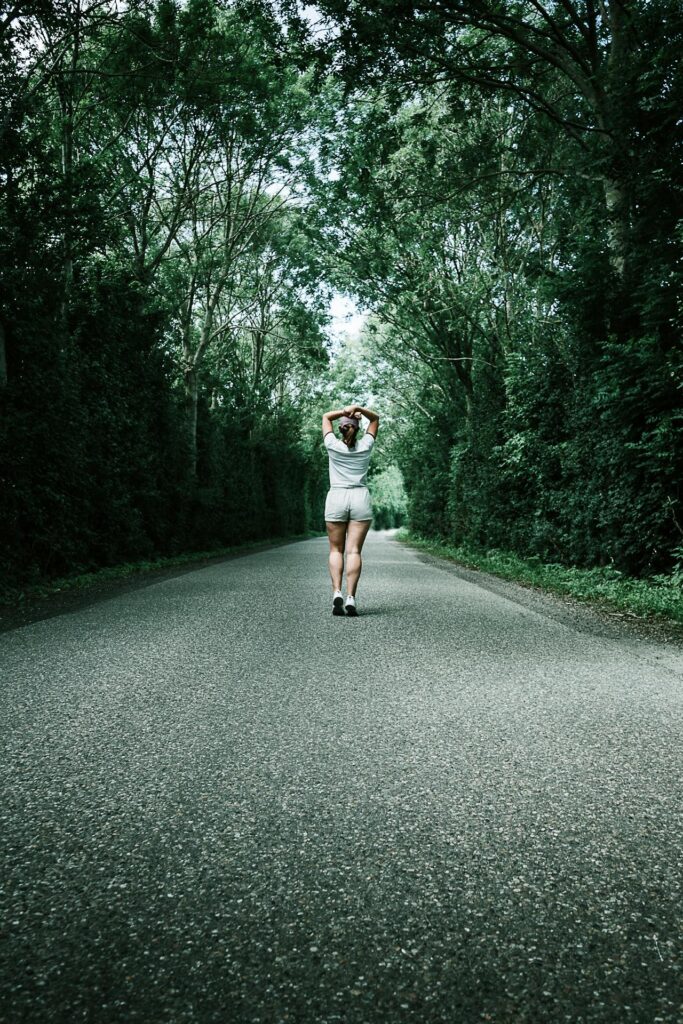 Image of a woman standing in the middle of a forest road, stretching arms behind their head under trees.