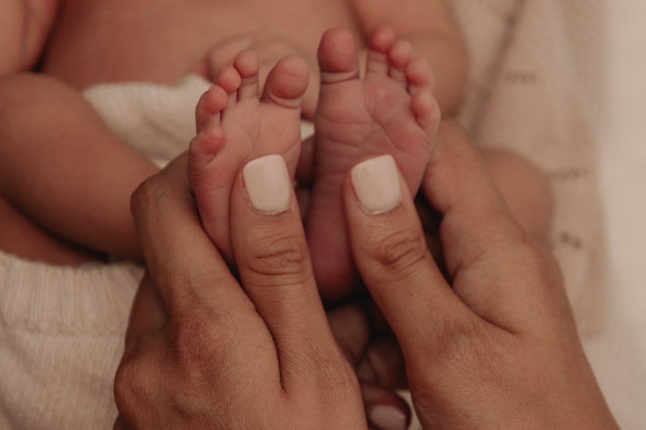 Close-up image of a newborn baby's tiny feet held gently by an adult's hands with neatly painted nails, showing I hate parenting.