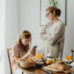 Image of a mother in a cardigan stands with arms crossed, looking at her daughter using a phone during breakfast, showing I hate parenting.