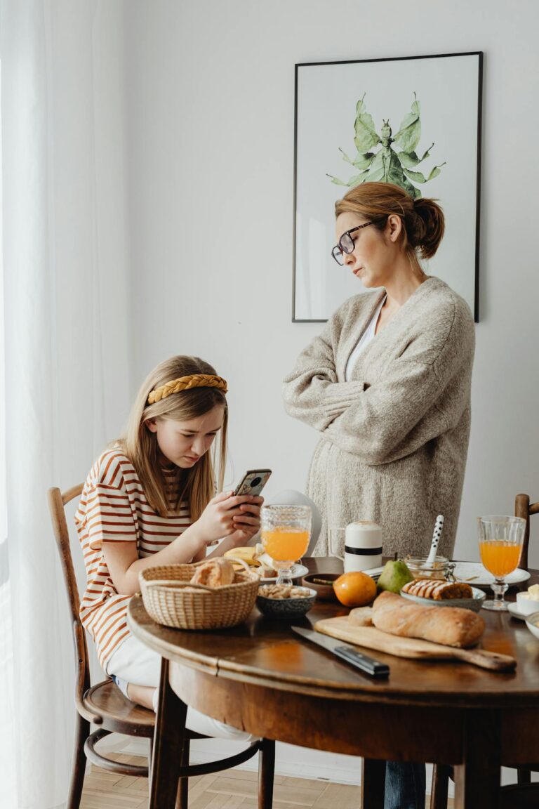 Image of a mother in a cardigan stands with arms crossed, looking at her daughter using a phone during breakfast, showing I hate parenting.