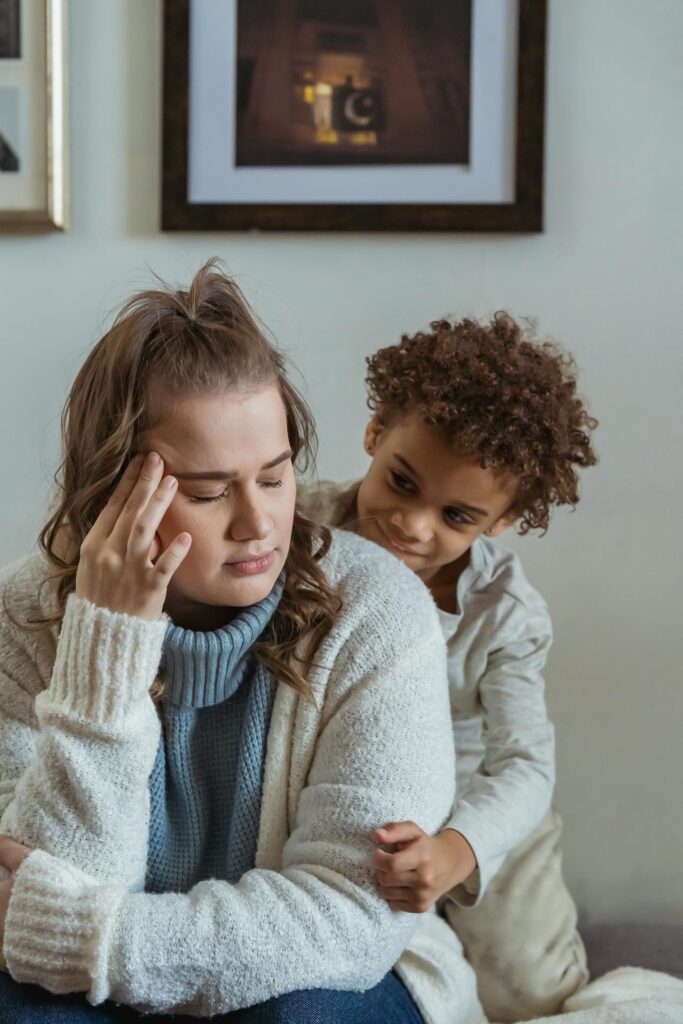 Image of a woman sitting with her head in her hand, while a child stands behind her, comforting her, representing stay-at-home mom syndrome.