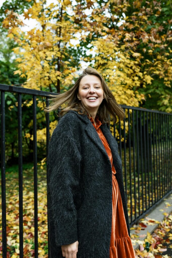 Image of a woman in a dark gray coat and orange dress, standing outdoors with autumn foliage behind, representing undergarments for plus-size women.