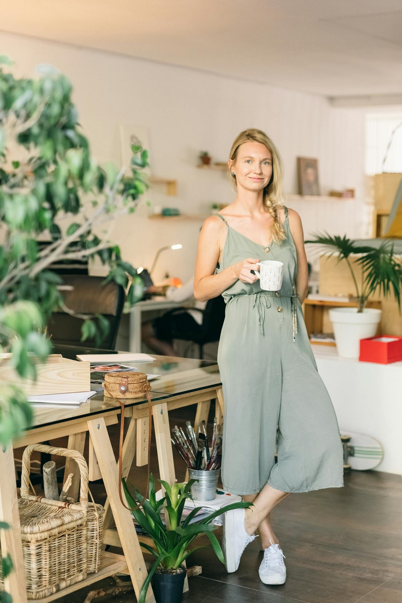 Image of a woman holding a mug, standing in an art studio with plants, wooden furniture, and painting supplies.