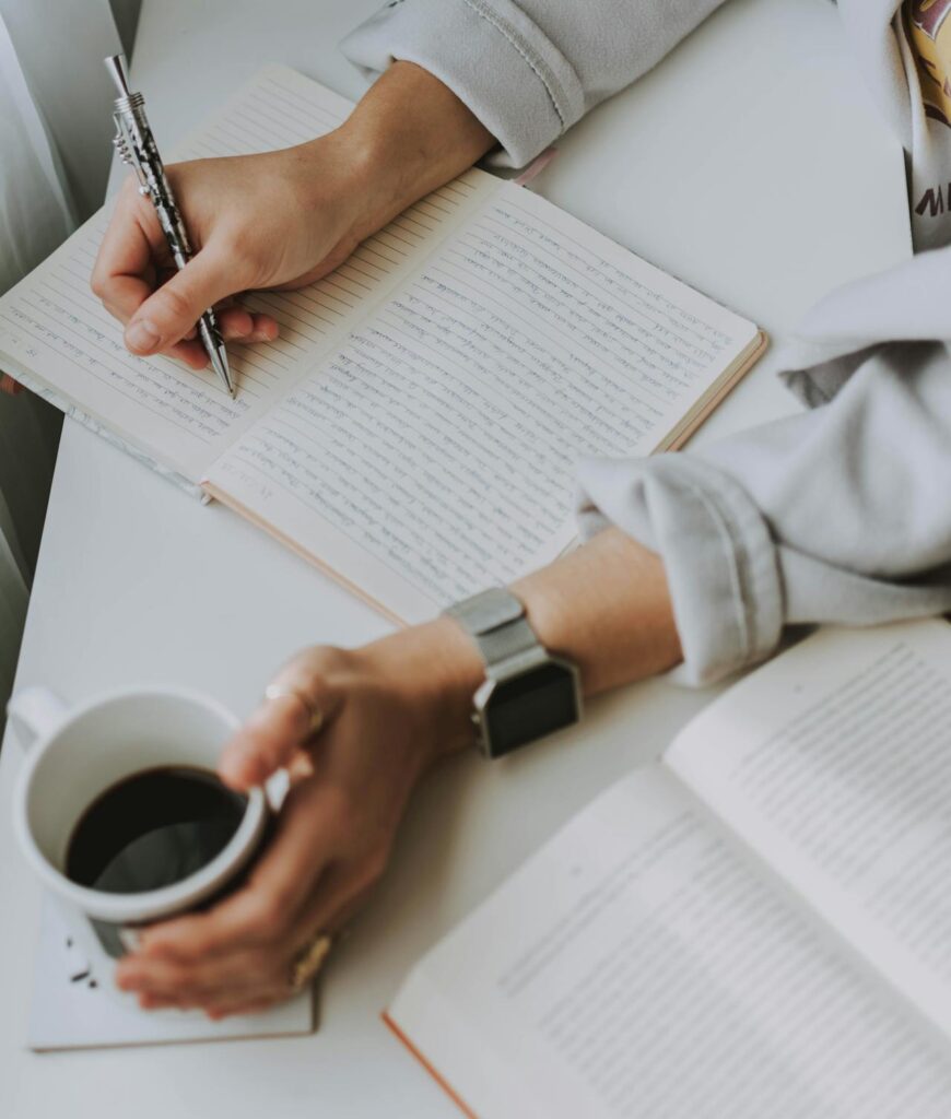 Close-up of hands writing in a journal while holding a coffee cup, with an open book nearby.