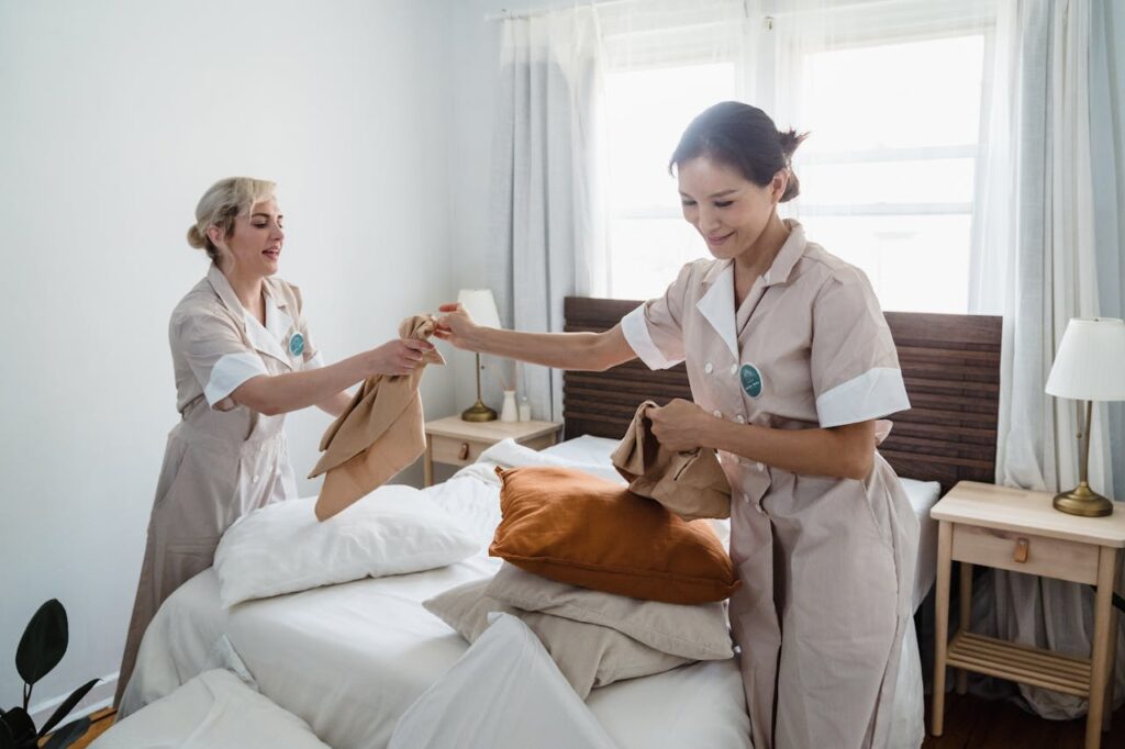 Image of two women dressed as hotel maids making a bed in a bright, tidy room with natural light, representing hire house help.