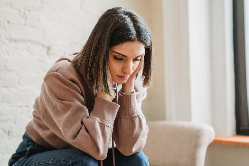 Image of a young woman sitting indoors, looking pensive with her hands resting on her face in thought.