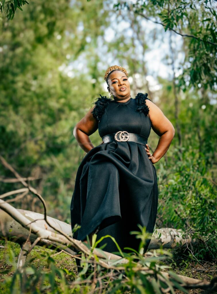 Image of a woman in a black dress with a golden crown poses confidently outdoors, surrounded by trees, representing  busy plus-size mom.