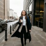 Image of a confident woman in a long coat walking on a city sidewalk holding a coffee cup, showing dress stylishly for family events as a mom.