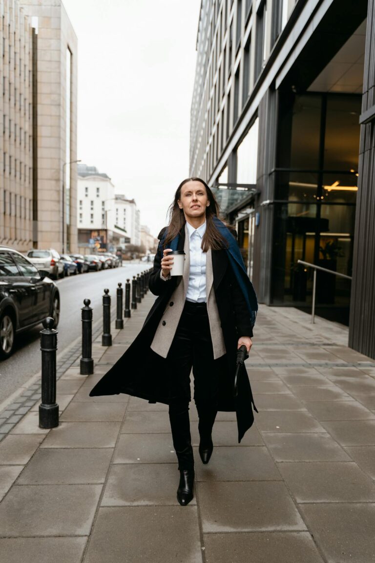 Image of a confident woman in a long coat walking on a city sidewalk holding a coffee cup, showing dress stylishly for family events as a mom.