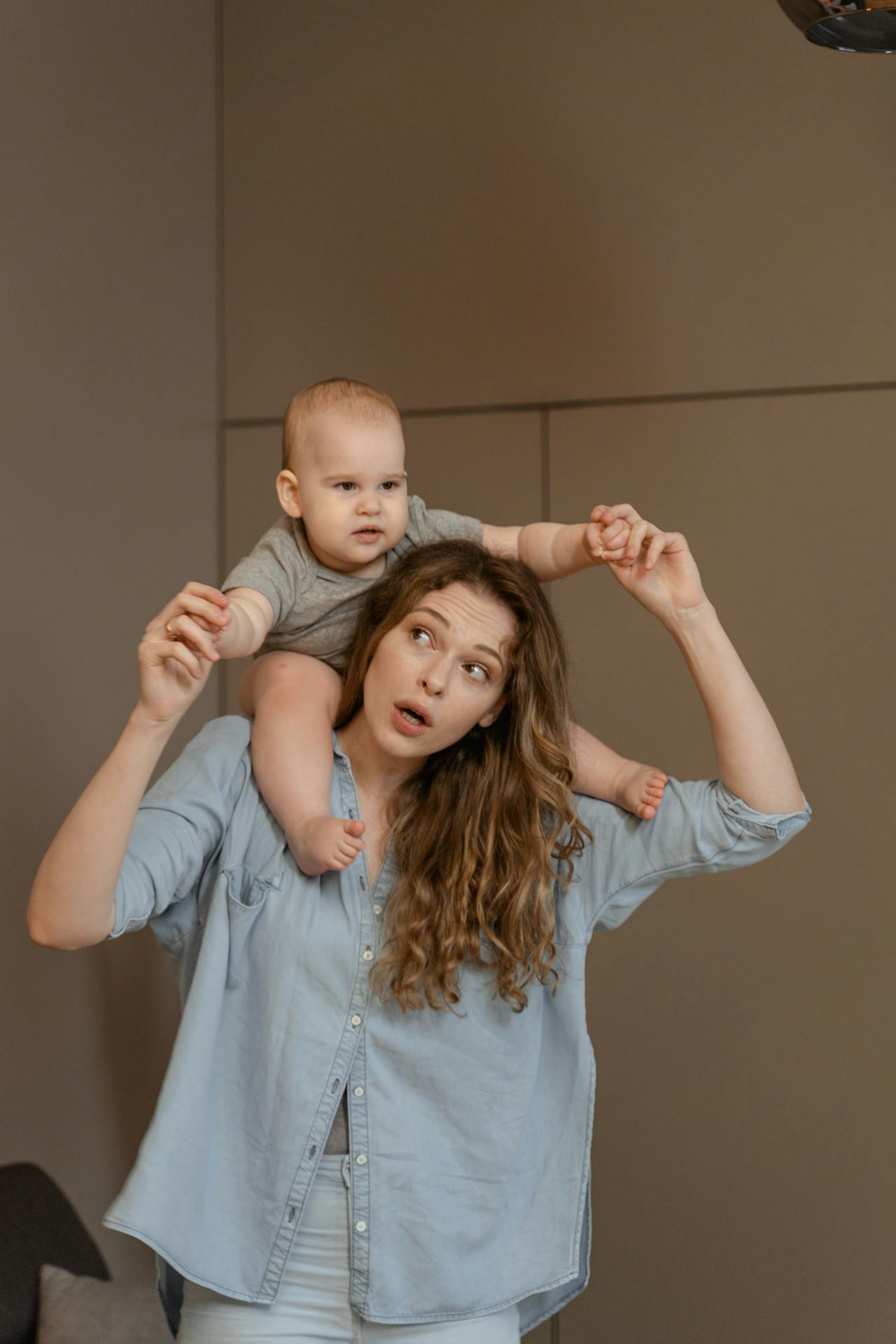 Close-up image of playful woman carrying a baby on her shoulders, wearing a casual blue shirt in a cozy room, showing best nursing outfits.