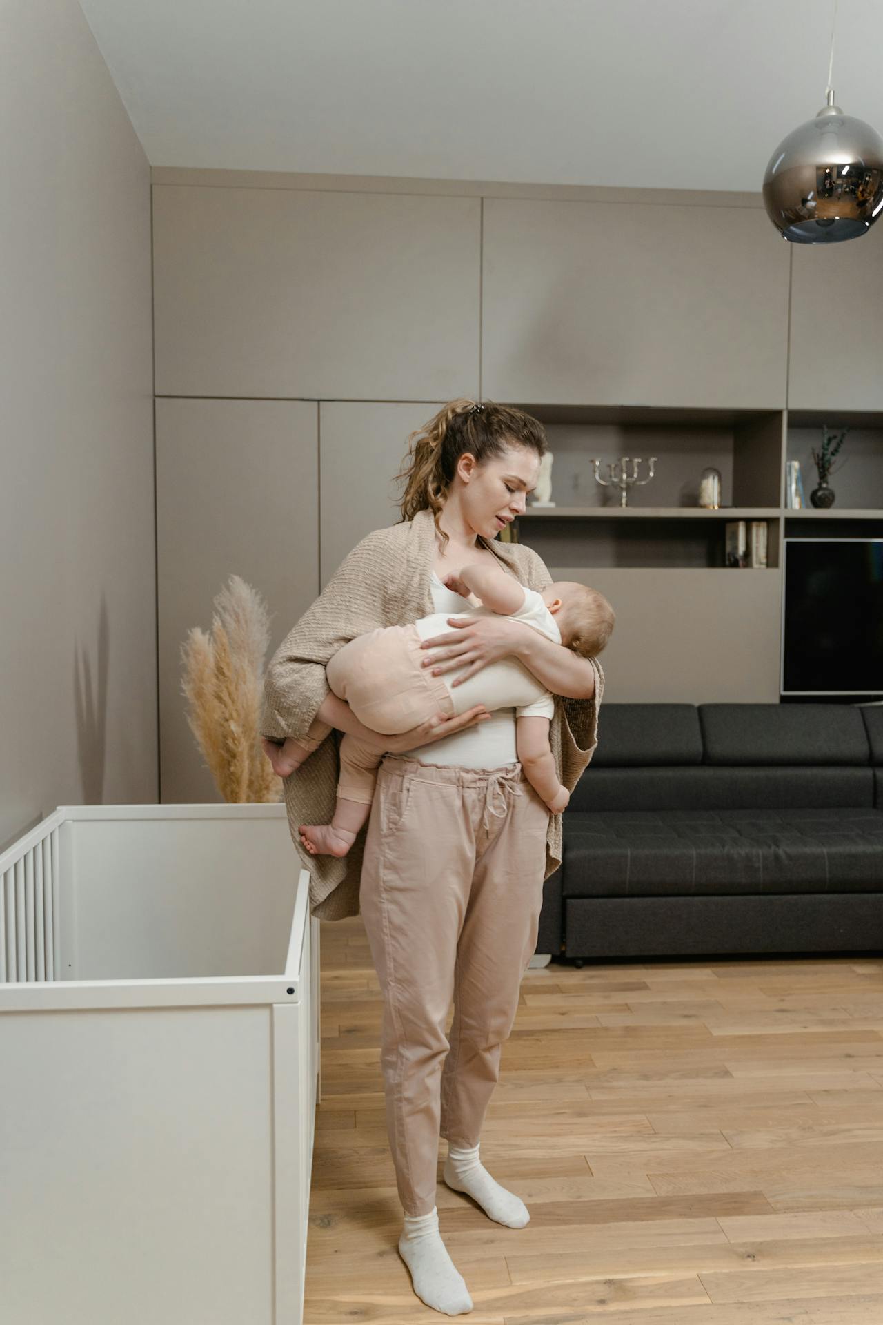 Image of a woman holding a baby near a white crib in a modern living room with wooden flooring, showing postpartum clothing.