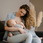 Image of a mother sits on a couch, breastfeeding her baby lovingly, with pampas grass decor in the background, showing postpartum outfits for breastfeeding mom.