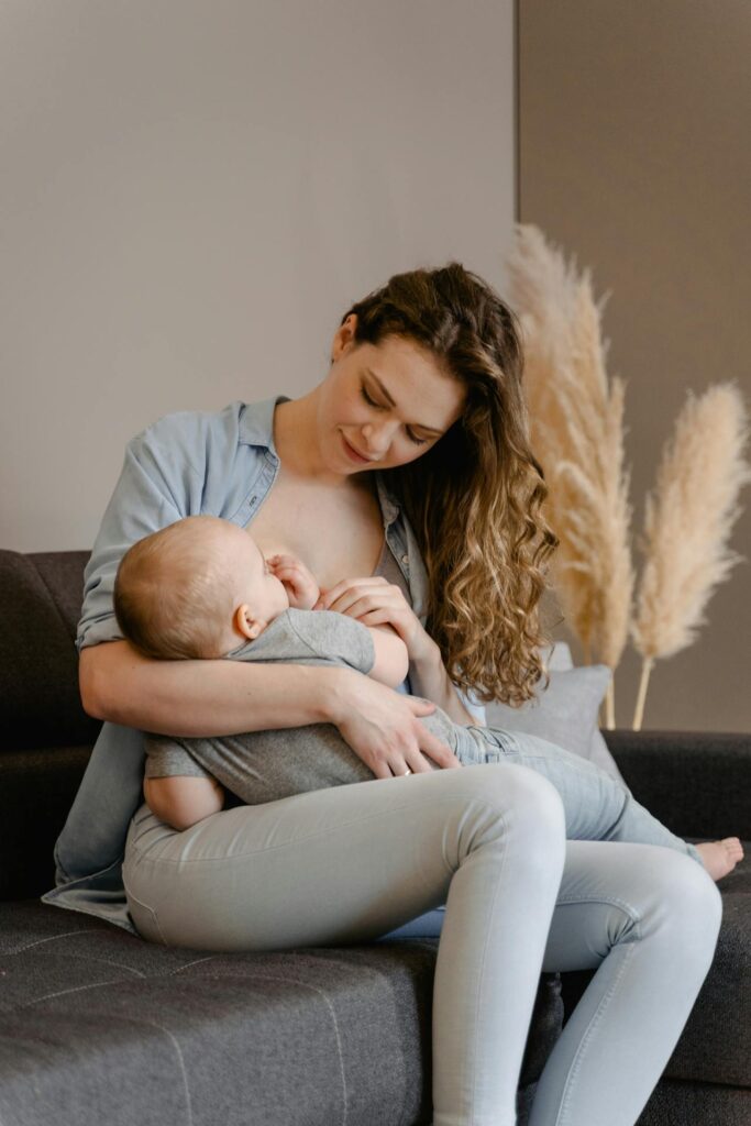 Image of a mother sits on a couch, breastfeeding her baby lovingly, with pampas grass decor in the background, showing postpartum outfits for breastfeeding mom.