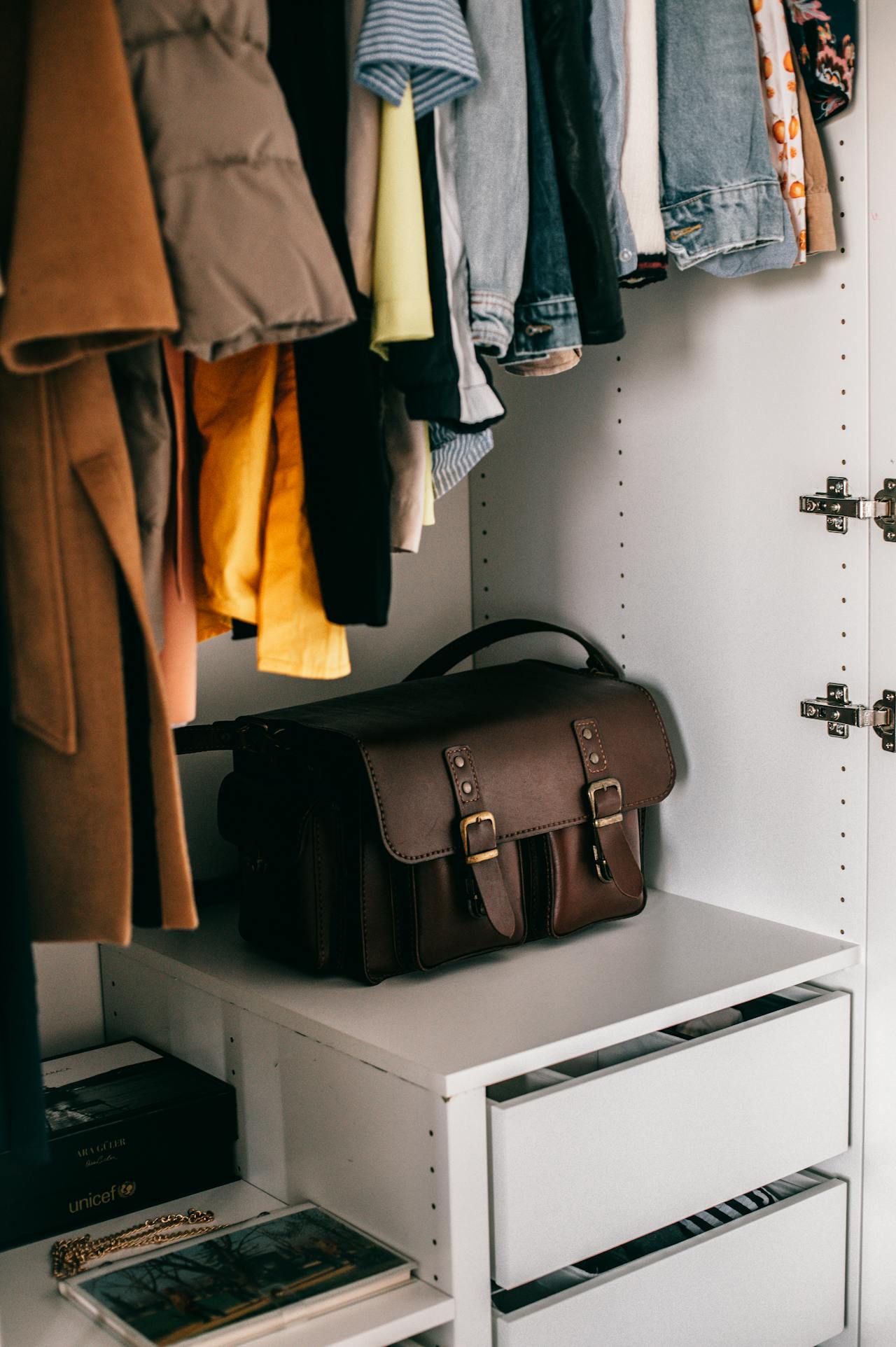 Image of a cozy wardrobe with colorful hanging clothes, a leather bag on a shelf, and drawers for storage.