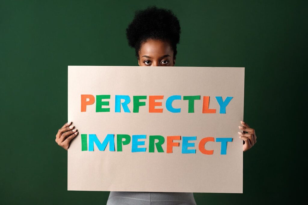 Image of a woman holding a sign that says, Perfectly Imperfect, in colorful letters on a green background.