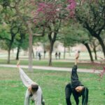 Image of two women practicing yoga outdoors in a park under a blooming tree with pink flowers, showing exercise after normal delivery to reduce tummy.