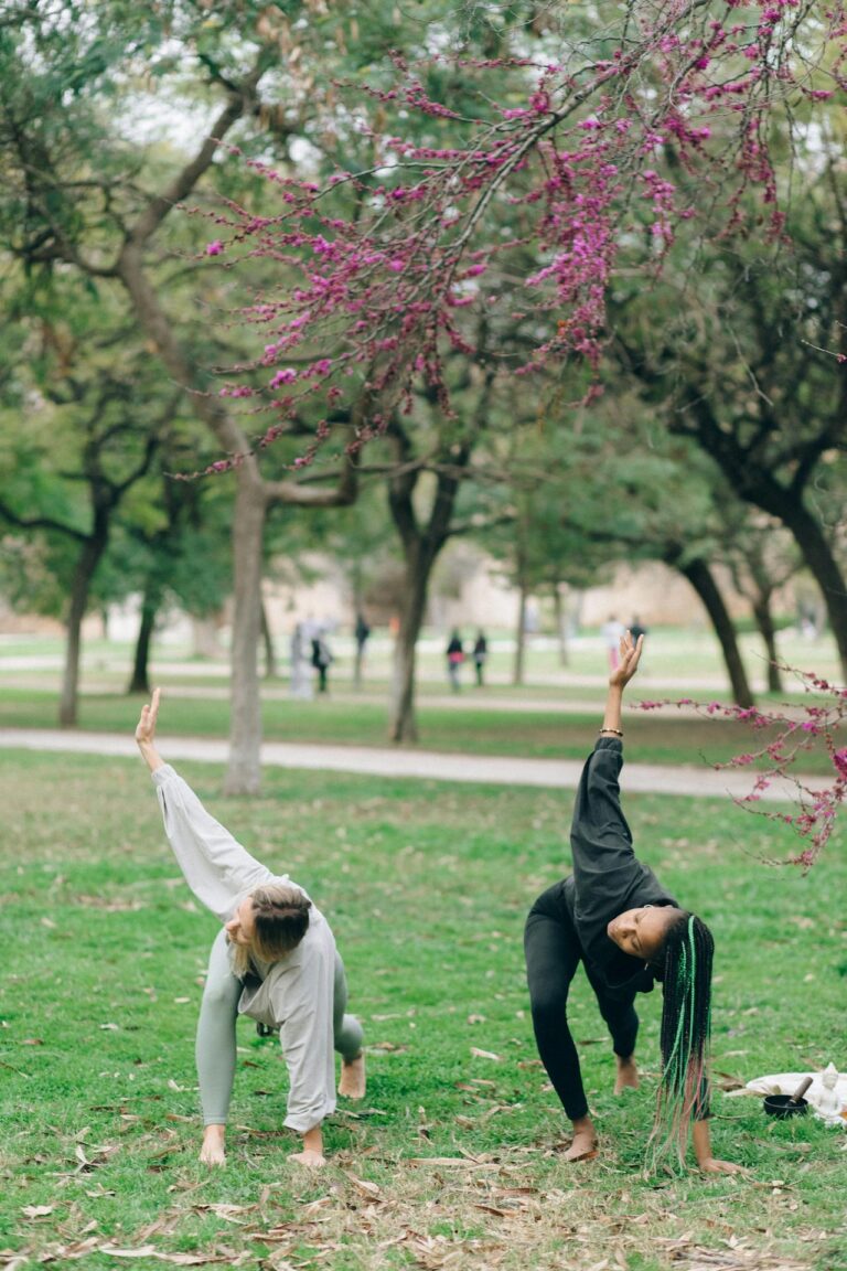 Image of two women practicing yoga outdoors in a park under a blooming tree with pink flowers, showing exercise after normal delivery to reduce tummy.