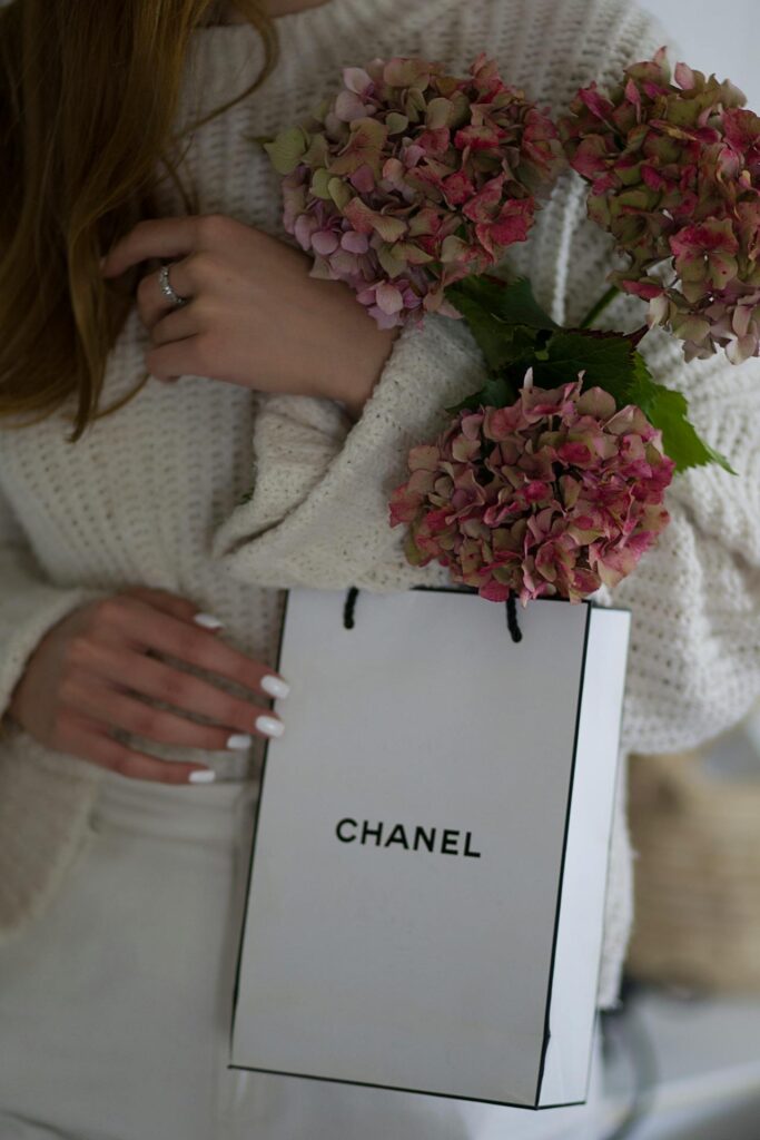 Image of a woman in a cozy white sweater holds pink hydrangea flowers and a Chanel shopping bag, showing build an eco-friendly wardrobe as a mom.