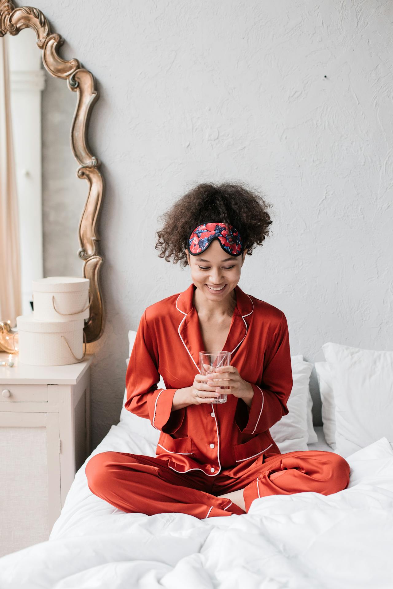 Image of a woman in red pajamas sitting on a bed, smiling while holding a glass of water with a sleep mask, showing pajama set for breastfeeding.