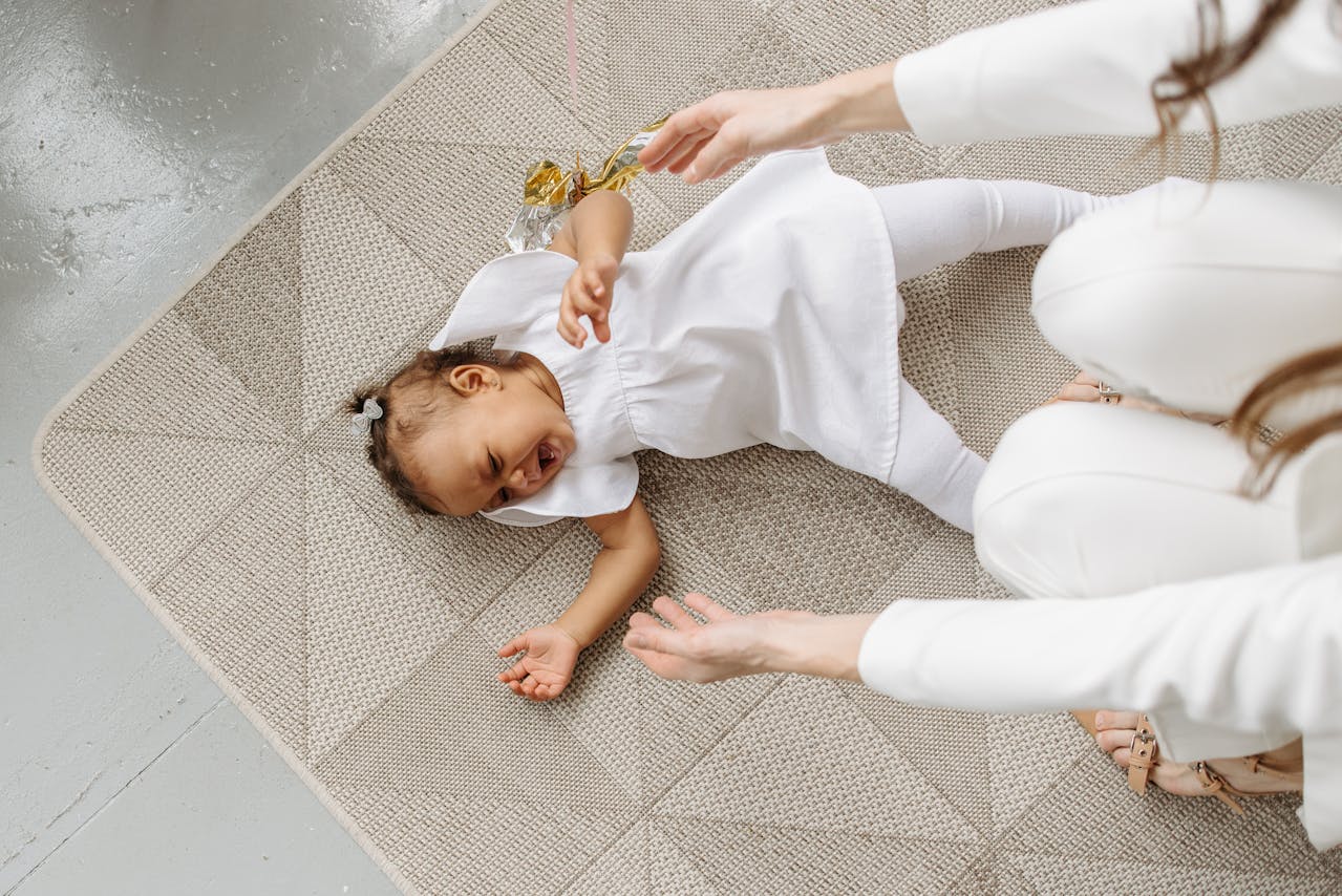 Image of a young child in a white dress crying on the floor as a woman reaches out to her.