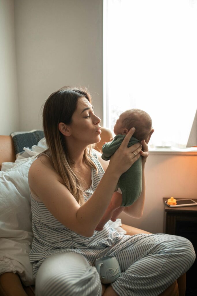 Image of a woman in striped pajamas holds a baby near a window, gently making a kissing gesture, representing exercise after normal delivery to reduce tummy.