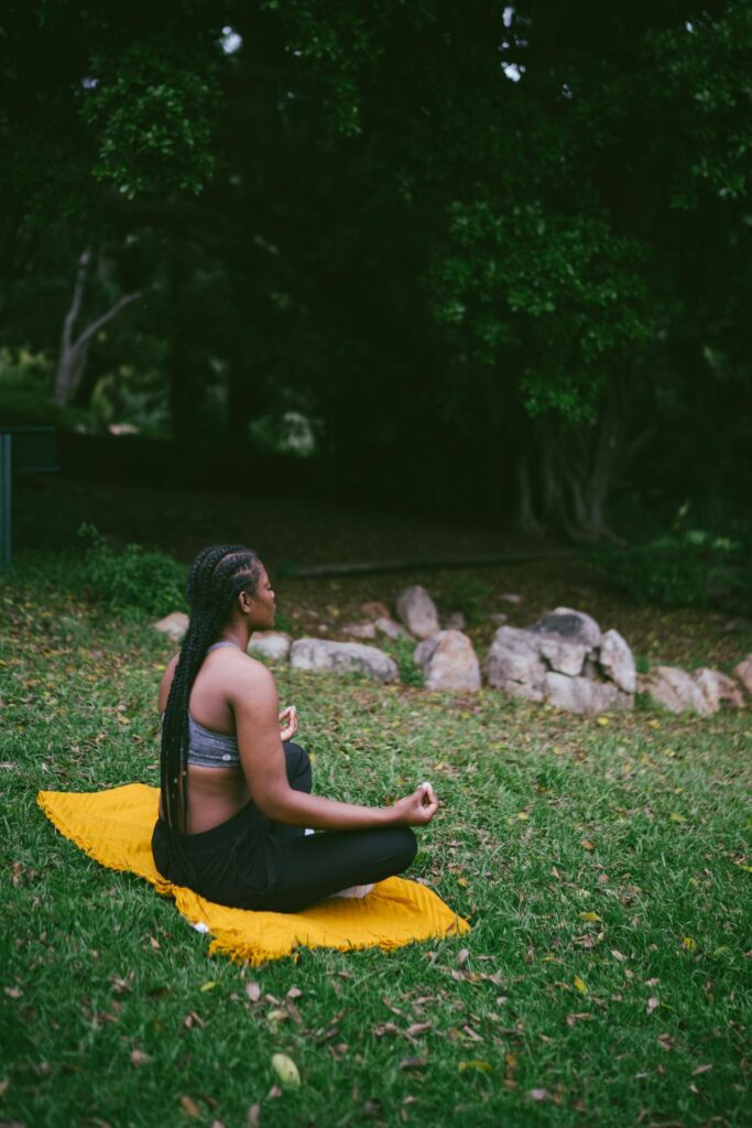 Image of a woman sits on a yellow mat meditating in a serene green outdoor space surrounded by trees.